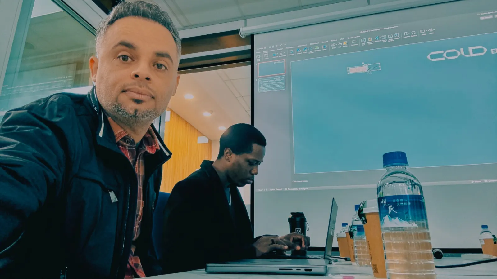 A selfie at the meeting table with a colleague on a laptop and the company deck on the projector
