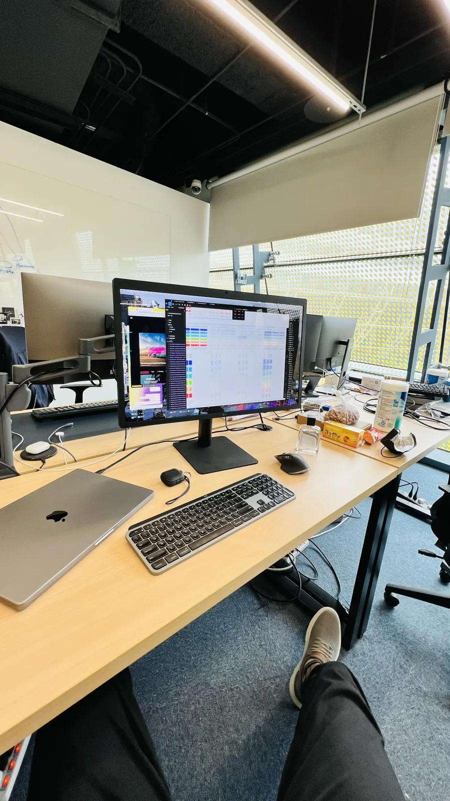 A desk by the window with a monitor, laptop, keyboard and snacks