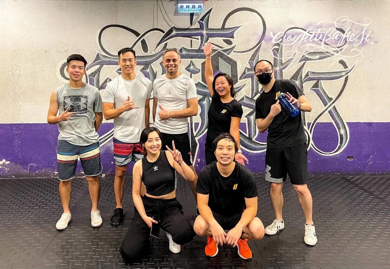 Seven people posing after a class in front of a graffiti wall, flashing peace signs and shakas